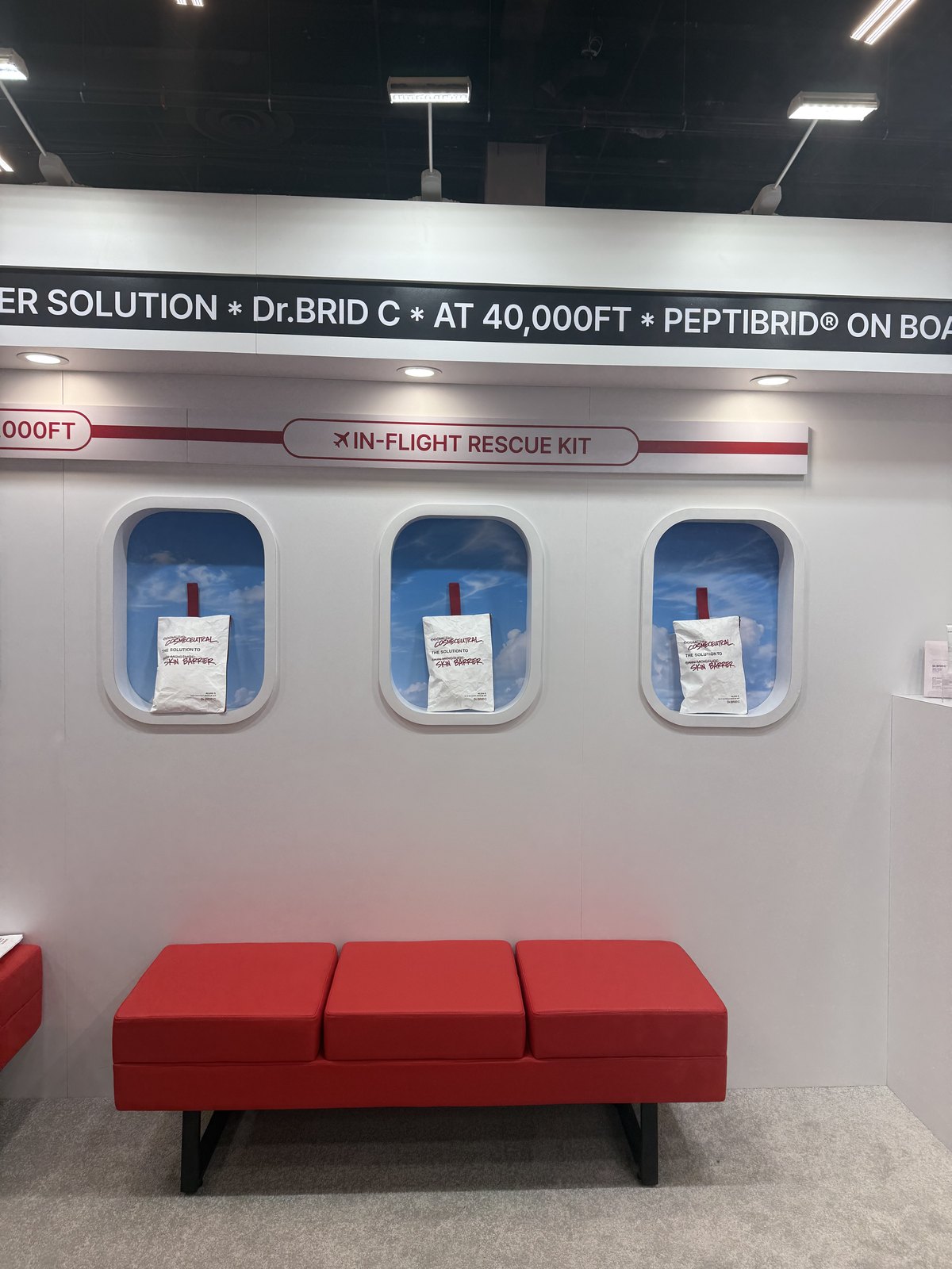 Aircraft windows cut into a white trade show wall, each holding a white sachet labelled in-flight rescue kit